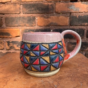 May include: A pink and white ceramic mug with a geometric pattern in shades of blue, red, and yellow. The mug has a handle that is painted orange.