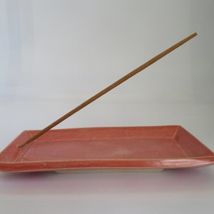 May include: A pink ceramic incense holder with a wooden incense stick resting on top.