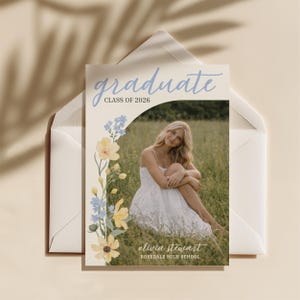 May include: A graduation announcement card with the word "graduate" in light blue script. The card features a photo of a person in a white dress, surrounded by a floral design with yellow and blue flowers. The text "Class of 2026" and the graduate's name and high school are also visible.