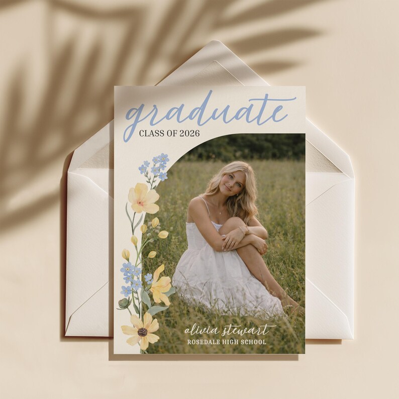 May include: A graduation announcement card with the word "graduate" in light blue script. The card features a photo of a person in a white dress, surrounded by a floral design with yellow and blue flowers. The text "Class of 2026" and the graduate's name and high school are also visible.