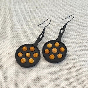 May include: A pair of black, pan-shaped earrings, each with a handle and filled with small, round, golden-brown objects. The earrings hang from black hooks.