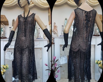May include: A black beaded flapper dress on a mannequin. The sleeveless dress features a scalloped hem and intricate beadwork. The mannequin also wears a beaded headband, long black gloves, and carries a small, beaded purse. The dress is a vintage style.