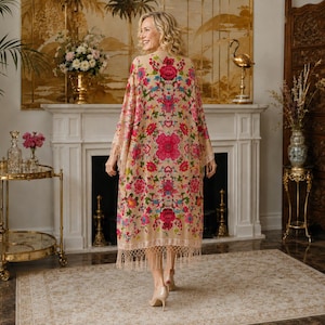 May include: A long, beige kimono-style robe featuring a vibrant, symmetrical floral pattern in shades of hot pink, red, turquoise, and orange. The garment has wide, flowing sleeves and a hemline finished with long, matching beige fringe trim.
