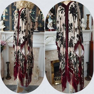 May include: A long, open-front kimono-style garment with a floral pattern in cream, black, and green. The edges and trim are a deep burgundy velvet, and the bottom features a fringe. The garment is displayed on a mannequin.