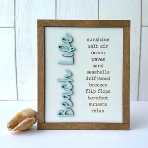 May include: A wooden framed sign with the words "Beach Life" in light blue script. The sign lists beach-related words like "sunshine," "sand," and "seashells." A seashell rests in front of the sign.