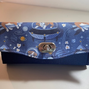 May include: A navy blue glasses case with a metal clasp. The top flap has a space-themed design with dogs in astronaut suits, planets, and stars. The base of the case is a solid navy blue.