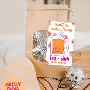 May include: A brown paper bag with a window showing silver disco balls. A pink and orange gift tag with a cartoon illustration of a mason jar filled with orange juice and a straw. The tag says "Thanks for being a sweet Tea-cher" and "From: Emmett".