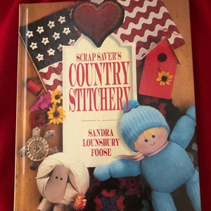 May include: Book cover with the title "Scrap Saver's Country Stitchery" by Sandra Lounsbury Foose. The cover features a variety of colorful fabric scraps, including a red heart, a red and white striped fabric, a red birdhouse, and a blue knitted doll.