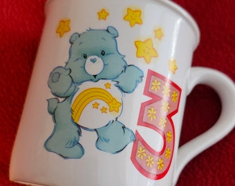May include: White ceramic mug with a blue Care Bear with a rainbow and stars on its chest. The Care Bear is holding a pink number 3 with yellow flowers on it. The mug is decorated with yellow stars.