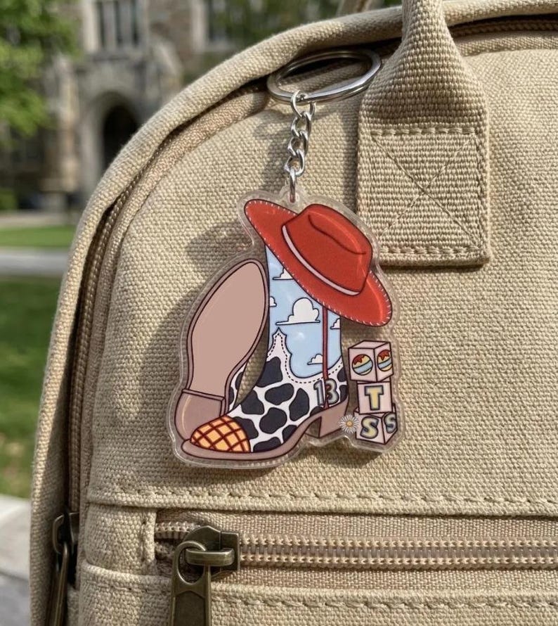 May include: A clear acrylic keychain featuring a cowboy boot with a cow print pattern, a blue sky with white clouds, and the number 13. A red cowboy hat rests on top of the boot, and a stack of three alphabet blocks with the letters T, S, and S sits nearby.