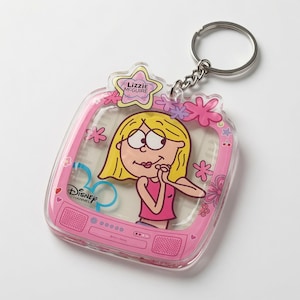 May include: A clear acrylic keychain shaped like a television set with a pink frame. The screen features a cartoon illustration of Lizzie McGuire with blonde hair and a pink top. The Disney Channel logo is printed on the left, and a star-shaped tag at the top reads Lizzie McGuire.