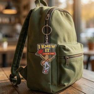 May include: An olive green canvas backpack with a clear acrylic keychain attached to the zipper. The keychain features a graphic of a person holding a red hat and guitar, with the yellow and red text I KNEW IT I KNEW YOU printed above.