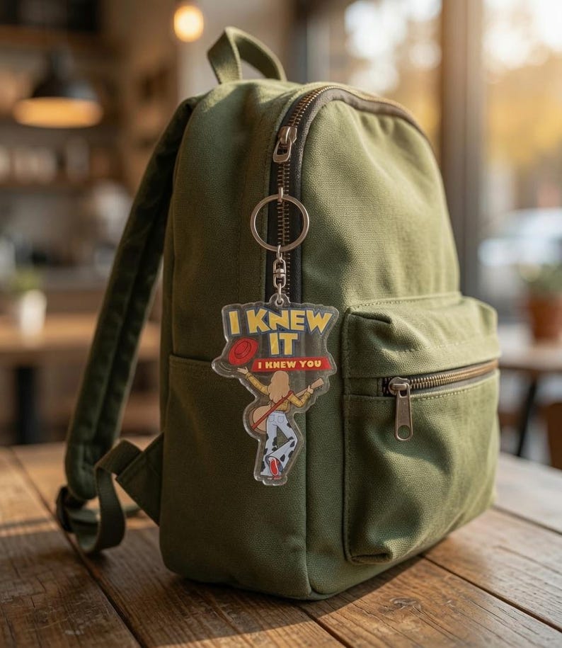 May include: An olive green canvas backpack with a clear acrylic keychain attached to the zipper. The keychain features a graphic of a person holding a red hat and guitar, with the yellow and red text I KNEW IT I KNEW YOU printed above.