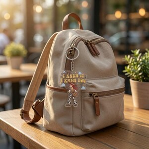 Puede incluir: Una mochila de lona beige con detalles en cuero marr&oacute;n tiene un llavero de acr&iacute;lico transparente colgado de la cremallera. El llavero muestra un personaje de dibujos animados tocando la guitarra con el texto I KNEW IT I KNEW YOU.