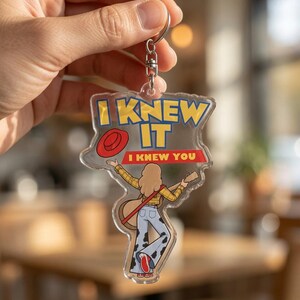 May include: A clear acrylic keychain featuring a cartoon illustration of a person with long hair wearing a yellow plaid shirt and cow-print bell-bottom pants, holding a guitar. The text reads I KNEW IT and I KNEW YOU, with a red hat icon.