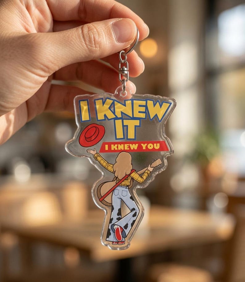 May include: A clear acrylic keychain featuring a cartoon illustration of a person with long hair wearing a yellow plaid shirt and cow-print bell-bottom pants, holding a guitar. The text reads I KNEW IT and I KNEW YOU, with a red hat icon.