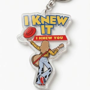 May include: A clear acrylic keychain featuring a cartoon illustration of a person with long hair wearing a yellow plaid shirt and cow-print bell-bottom pants, holding a guitar. The text reads I KNEW IT and I KNEW YOU, with a red hat graphic.
