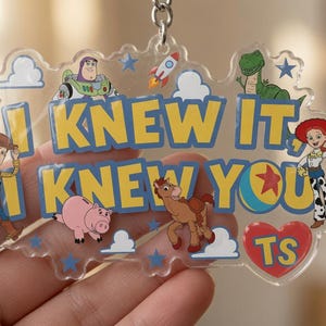 May include: A clear acrylic keyring featuring Toy Story characters including Woody, Buzz Lightyear, Rex, Jessie, Bullseye, and Hamm. The text reads I KNEW IT, I KNEW YOU with a red heart containing the letters TS. Blue stars and white clouds decorate the piece.