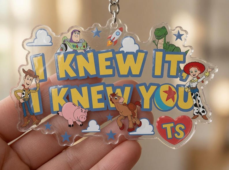 May include: A clear acrylic keyring featuring Toy Story characters including Woody, Buzz Lightyear, Rex, Jessie, Bullseye, and Hamm. The text reads I KNEW IT, I KNEW YOU with a red heart containing the letters TS. Blue stars and white clouds decorate the piece.