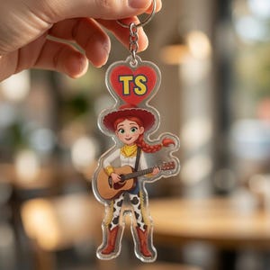 May include: A clear acrylic keyring featuring a cartoon character with red hair in a braid, wearing a red cowboy hat, yellow neckerchief, and cow-print chaps, playing an acoustic guitar. Above the character is a red heart with the yellow text TS.