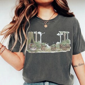 May include: Dark grey t-shirt featuring a cartoon illustration of Grogu in a forest setting. The design includes Grogu in a pod and several Grogu figures with the word "cool" in the background. The shirt is made of a soft material.