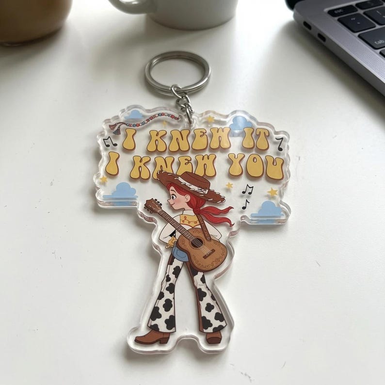 Puede incluir: Un llavero de acr&iacute;lico transparente con una vaquera de dibujos animados de pelo rojo, sombrero marr&oacute;n y pantalones con estampado de vaca tocando una guitarra ac&uacute;stica. El texto "I KNEW IT I KNEW YOU" aparece en letras amarillas sobre la figura.
