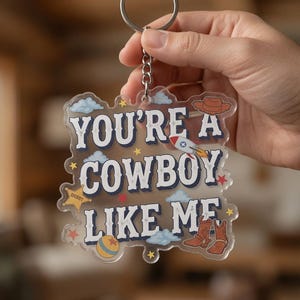 May include: A clear acrylic keychain featuring the text "YOU'RE A COWBOY LIKE ME" in dark blue lettering. The design includes illustrations of a brown cowboy hat, a rocket ship, a yellow ball, brown cowboy boots, blue clouds, and small yellow stars.