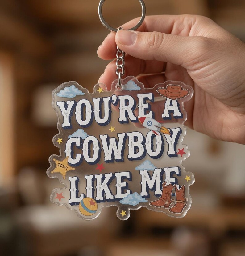 May include: A clear acrylic keychain featuring the text "YOU'RE A COWBOY LIKE ME" in dark blue lettering. The design includes illustrations of a brown cowboy hat, a rocket ship, a yellow ball, brown cowboy boots, blue clouds, and small yellow stars.
