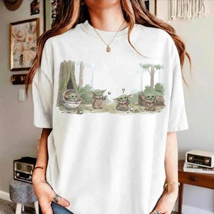 May include: White t-shirt featuring a cartoon illustration of Grogu from The Mandalorian. The design shows Grogu in various poses within a forest setting. The word "Cool" is written in a speech bubble. A fun, casual tee.