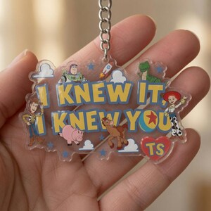 May include: A clear acrylic keyring featuring the yellow text I KNEW IT, I KNEW YOU with illustrations of Woody, Buzz Lightyear, Rex, Jessie, Bullseye, and Hamm from Toy Story, along with a red heart containing the letters TS and a metal chain.