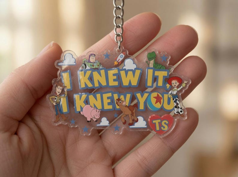 May include: A clear acrylic keyring featuring the yellow text I KNEW IT, I KNEW YOU with illustrations of Woody, Buzz Lightyear, Rex, Jessie, Bullseye, and Hamm from Toy Story, along with a red heart containing the letters TS and a metal chain.