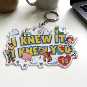 May include: A clear acrylic keyring featuring the text I KNEW IT, I KNEW YOU in yellow letters. The design includes colourful illustrations of cartoon characters, a rocket, blue stars, white clouds, and a red heart with the letters TS inside.