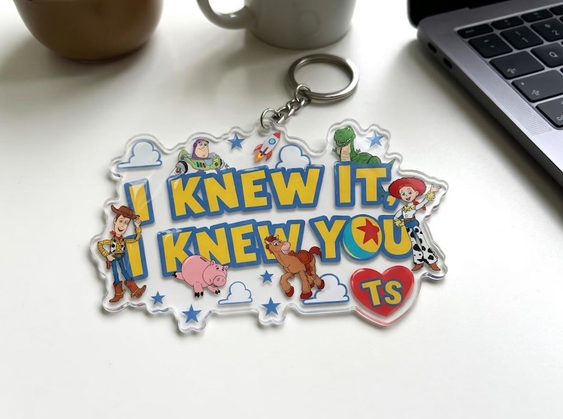May include: A clear acrylic keyring featuring the text I KNEW IT, I KNEW YOU in yellow letters. The design includes colourful illustrations of cartoon characters, a rocket, blue stars, white clouds, and a red heart with the letters TS inside.