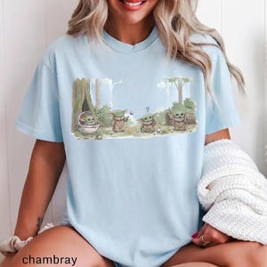 May include: Light blue t-shirt featuring a cartoon illustration of a character from a popular science fiction series. The graphic shows the character in various poses within a forest setting. The word "cool" is written on the graphic.