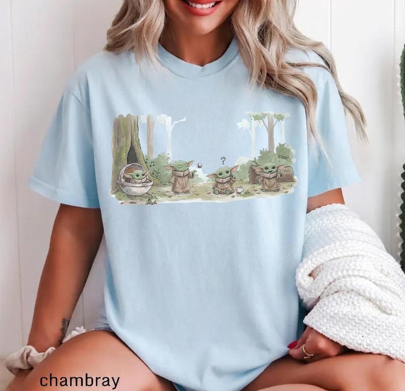 May include: Light blue t-shirt featuring a cartoon illustration of a character from a popular science fiction series. The graphic shows the character in various poses within a forest setting. The word "cool" is written on the graphic.