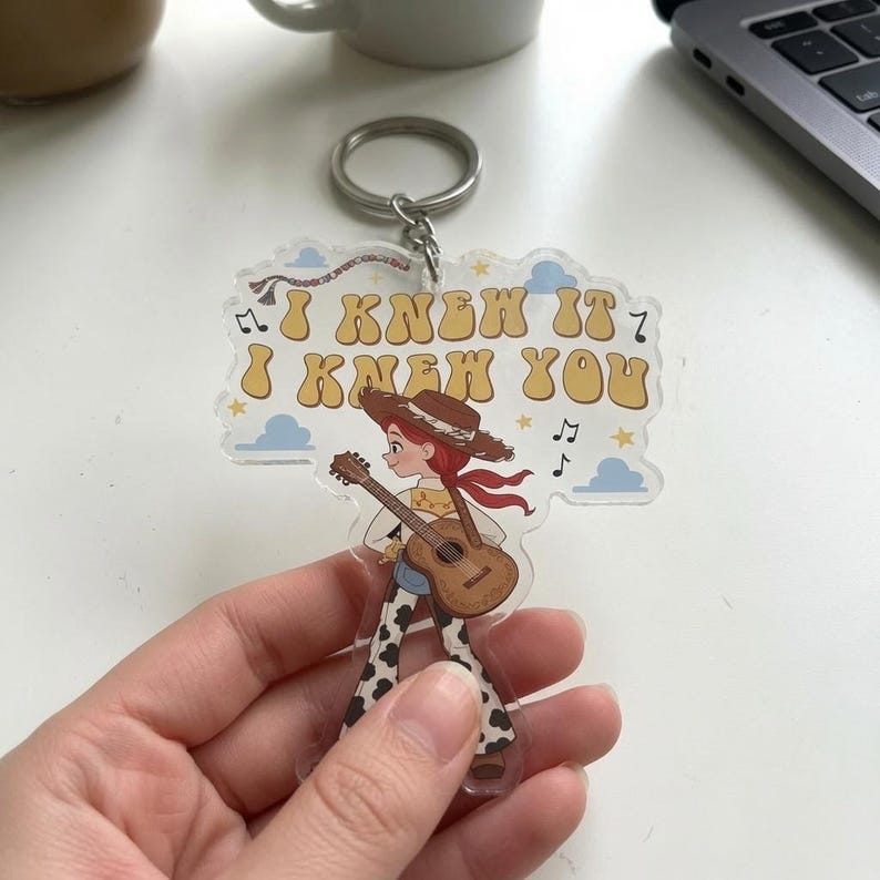 Puede incluir: Un llavero de acr&iacute;lico transparente con una vaquera de dibujos animados de pelo rojo, sombrero marr&oacute;n y pantalones con estampado de vaca tocando la guitarra. El texto "I KNEW IT I KNEW YOU" est&aacute; impreso en amarillo con borde marr&oacute;n y nubes azules.