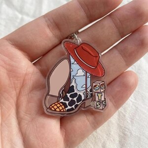 May include: An acrylic keychain featuring a cowboy boot with a cow print pattern, a blue sky and cloud design, and the number 13. A red cowboy hat rests on the boot, and three stacked blocks are visible with the letters T, S, and S printed on them.