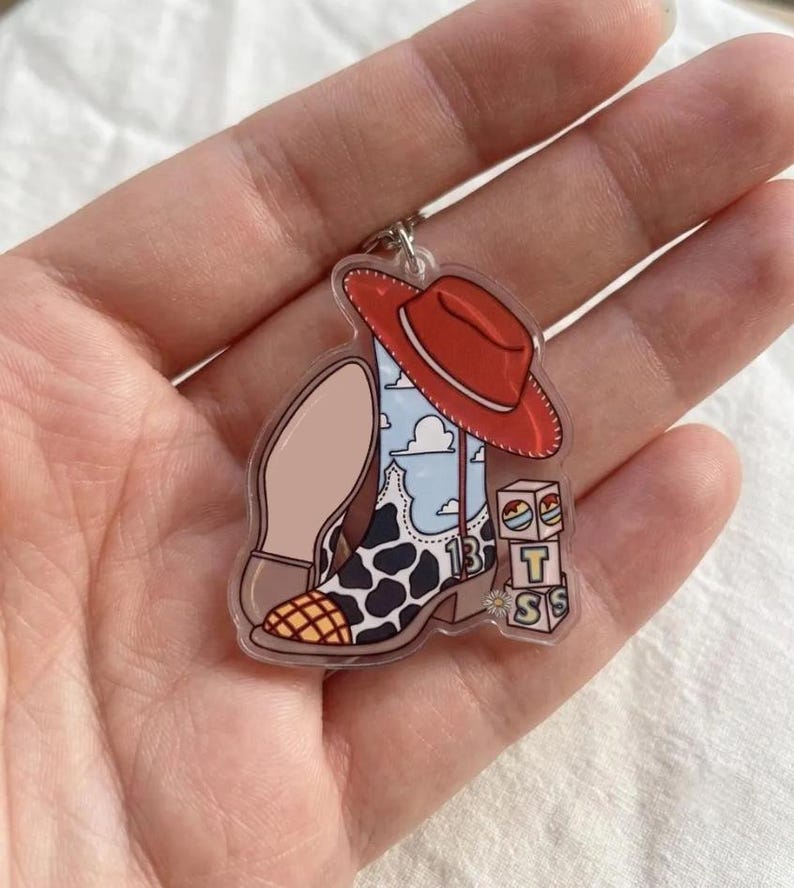 May include: An acrylic keychain featuring a cowboy boot with a cow print pattern, a blue sky and cloud design, and the number 13. A red cowboy hat rests on the boot, and three stacked blocks are visible with the letters T, S, and S printed on them.