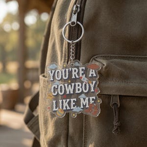 May include: A clear acrylic keychain featuring the text "YOU'RE A COWBOY LIKE ME" in dark blue letters. The design includes small illustrations of a rocket, a cowboy hat, cowboy boots, a ball, clouds, and stars. It hangs from a silver metal ring.