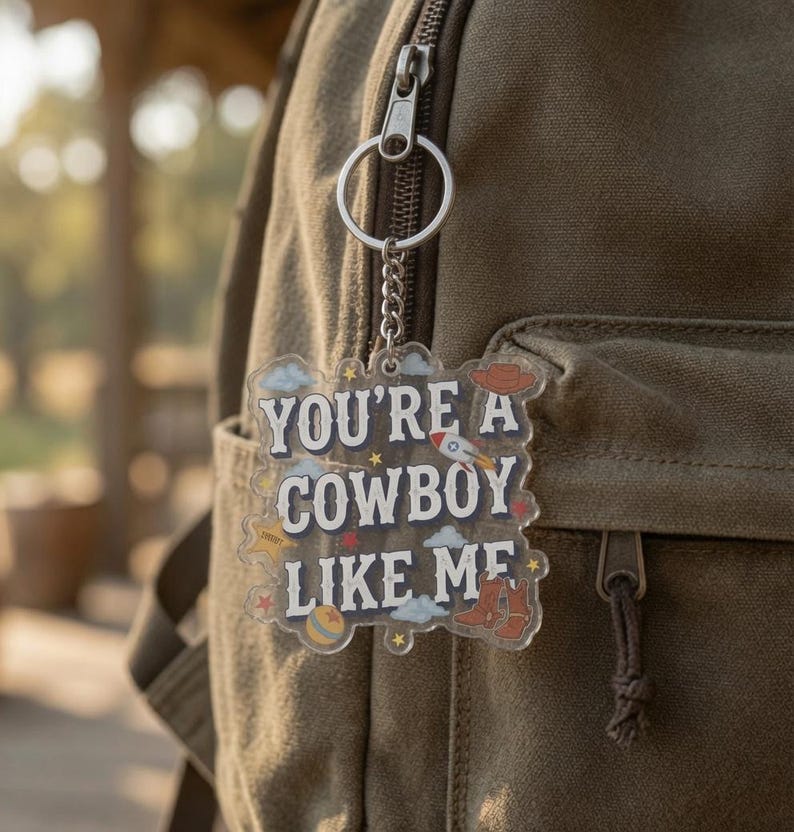 May include: A clear acrylic keychain featuring the text "YOU'RE A COWBOY LIKE ME" in dark blue letters. The design includes small illustrations of a rocket, a cowboy hat, cowboy boots, a ball, clouds, and stars. It hangs from a silver metal ring.