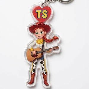 May include: An acrylic keyring featuring a cartoon cowgirl with red hair in a braid, wearing a red hat, white shirt, and cow-print trousers, playing an acoustic guitar. Above her is a red heart with the yellow letters TS printed inside.