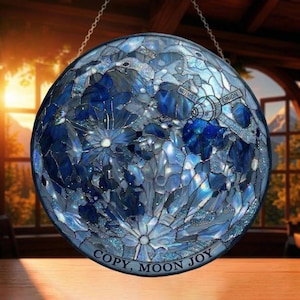 Op de afbeelding: Rond glas-in-lood kunstwerk dat de maan afbeeldt in blauwe en witte tinten. Het ontwerp bevat de tekst "COPY, MOON JOY" en afbeeldingen van de Artemis II en Orion ruimtevaartuigen. Opgehangen aan een ketting.
