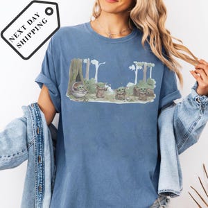 May include: Blue t-shirt featuring a graphic of Grogu from The Mandalorian in a forest scene. The design includes Grogu in a pod and standing with other characters. The shirt has the text "Cool" on the design.
