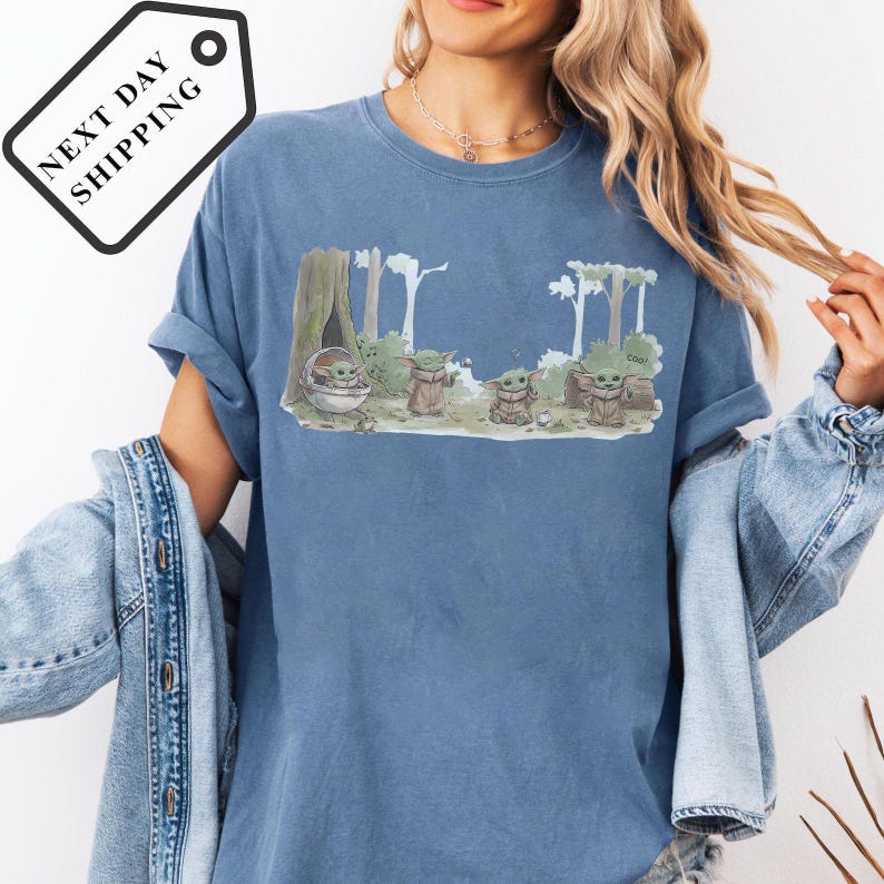 May include: Blue t-shirt featuring a graphic of Grogu from The Mandalorian in a forest scene. The design includes Grogu in a pod and standing with other characters. The shirt has the text "Cool" on the design.