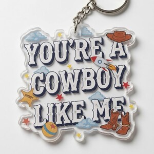 May include: A clear acrylic keychain featuring the text YOU'RE A COWBOY LIKE ME in dark blue letters. The design includes a brown cowboy hat, a rocket ship, a yellow sheriff star, a round ball, and brown cowboy boots with light blue clouds.