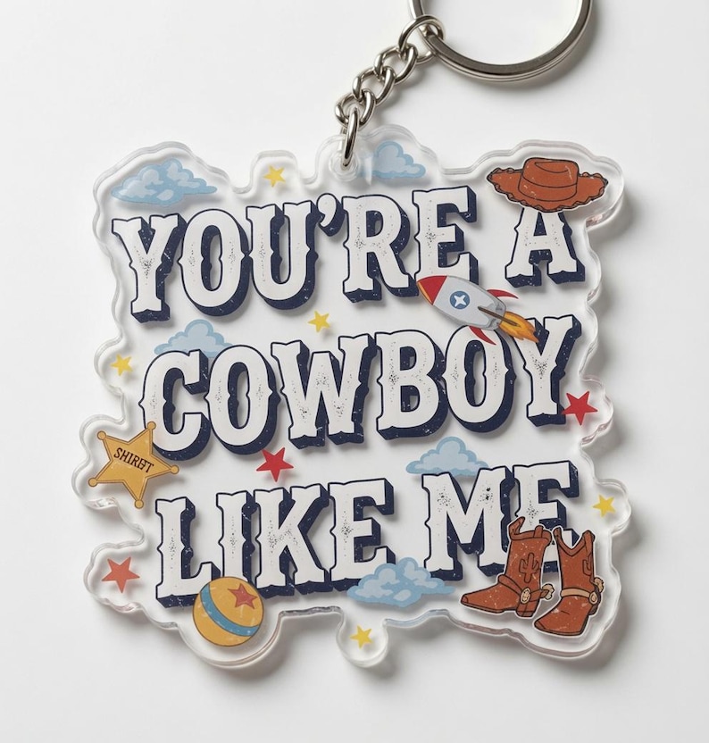 May include: A clear acrylic keychain featuring the text YOU'RE A COWBOY LIKE ME in dark blue letters. The design includes a brown cowboy hat, a rocket ship, a yellow sheriff star, a round ball, and brown cowboy boots with light blue clouds.