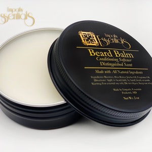 May include: A black tin with a silver lid containing a white beard balm. The tin has gold lettering that reads "Impala Essentials Beard Balm Conditioning Softener Distinguished Scent Made with All Natural Ingredients Made by Impala Essentials Frederick, MD Net Wt. 2 oz."