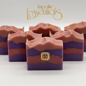 May include: Five bars of handmade soap with a layered design. The soap is a deep purple color with a thin layer of gold and a top layer of a light pink color. The soap is arranged in a row on a white surface. The text "Empathy Essentials" is visible in the top center of the image.