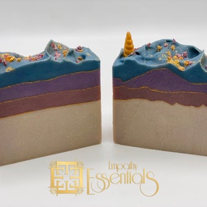 May include: Two bars of handmade soap with a unicorn horn on top. The soap is layered in shades of purple, blue, and beige with gold glitter and pink sprinkles on top. The soap is labeled "Empathy Essentials".