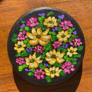 May include: A round canvas painting with a black background and a colorful floral design. The flowers are yellow, pink, and purple, and they are arranged in a cluster in the center of the canvas.